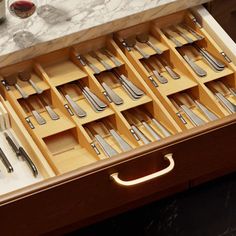 an open drawer with silverware in it on a marble counter top next to wine glasses and utensils