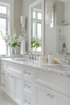 a white bathroom with double sinks and large mirrors on the wall next to two windows