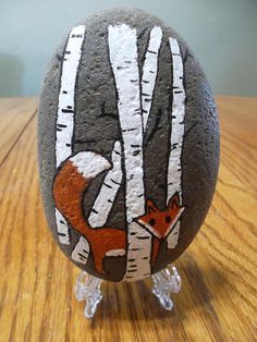 a rock painted with an image of a fox and birch trees, on a wooden table