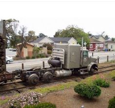 an old truck is parked on the tracks near some bushes and flowers in front of houses