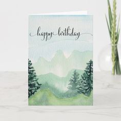 a card with the words happy birthday written in black ink on top of it, surrounded by pine trees and mountains