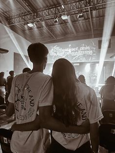 two people standing in front of a stage with their arms around each other as they look at the screen