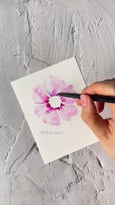 a hand holding a pen and drawing a flower on a piece of paper with watercolor pencils