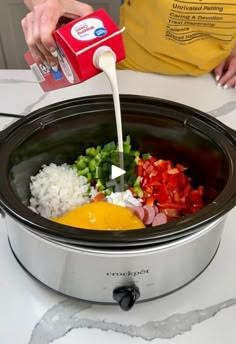 someone pouring milk into a crock pot filled with vegetables and rice on top of a table