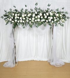 an arrangement of white flowers and greenery in front of a drapeed backdrop