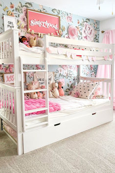 White solid wood bunk bed with trundle for kids. Girl's bedroom with floral wallpaper and pink decor. Toddler Girl Bunk Bed Ideas, Bunk Bed Rooms Girls Decor, Sister Bedroom Bunkbeds, White Bunk Beds Shared Bedrooms, Bunk Bed Decorating Ideas Diy, Girls Room With Bunk Beds Decor, Sister Bunk Beds Bedroom Ideas, Sister Bedroom Ideas Toddler