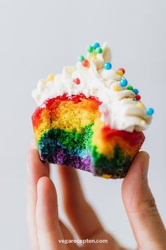 someone is holding up a rainbow cupcake with white frosting and sprinkles