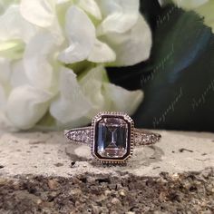 a fancy ring with an emerald colored stone surrounded by diamonds on a rock next to white flowers