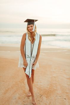 a woman standing on top of a sandy beach wearing a graduation cap and gown in front of the ocean