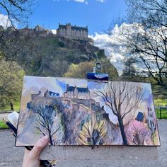 someone is holding up an art piece in front of a castle with trees and bushes