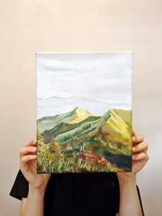 a woman holding up a painting with mountains in the background