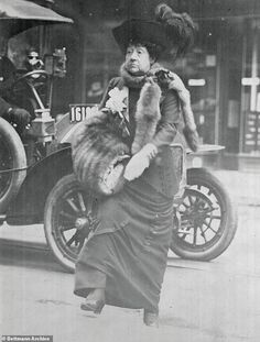 an old fashion photo of a woman in fur stoler and hat with car behind her