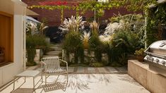 an outdoor patio with a grill, chairs and plants on the side walk in front of it