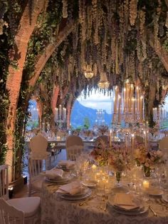 a table set up for a formal dinner with candles and flowers on the ceiling, surrounded by greenery