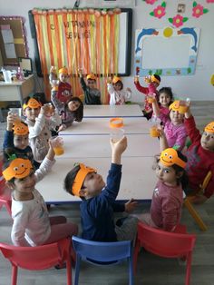 a group of children sitting at a table with orange hats on their heads and hands in the air