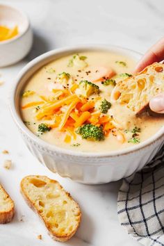 a person holding a piece of bread over a bowl of broccoli cheese soup