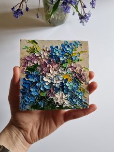 a hand holding up a piece of art with flowers in the back ground and on top of it