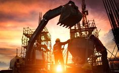 two construction workers standing next to each other in front of a large crane at sunset