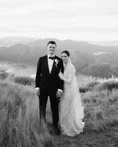 a bride and groom standing on top of a grass covered hill with mountains in the background