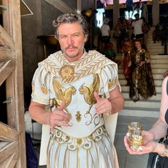 a man dressed in an ancient greek costume holding two gold cups while standing next to a woman