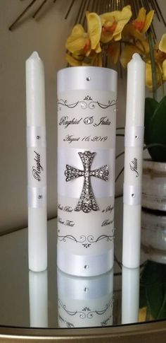 three white candles are sitting on a table with flowers in the background and one candle has a cross on it