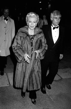 an older woman in a fur coat and tuxedo walking down the street with another man