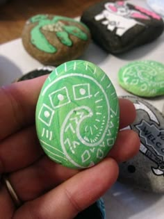 a hand holding a green and white painted button in front of several rocks with designs on them