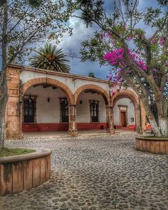 an old spanish style house with pink flowers on the trees and cobblestone walkway