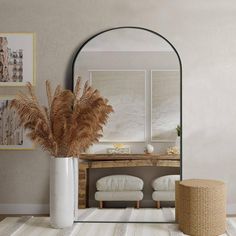 a white vase with dried plants in it next to a mirror on the wall near a footstool