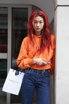 a woman with red hair and headphones leaning against a wall holding a shopping bag