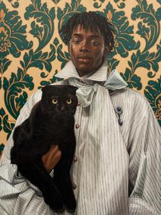 a man holding a black cat in front of a wall with green and gold damask