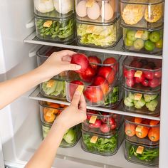 a person reaching for an apple in front of a refrigerator full of fruit and vegetables