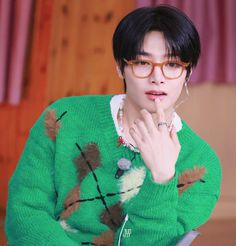 a young man wearing glasses and a green sweater is looking at the camera with his hand on his chin