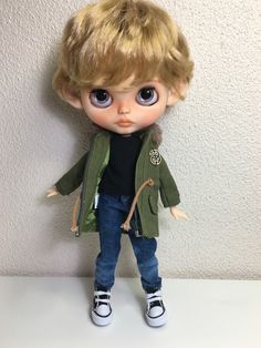 a doll with blonde hair and blue eyes is standing on a white surface, wearing jeans and a green jacket