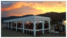 a group of people standing around in front of a white structure on the beach at sunset