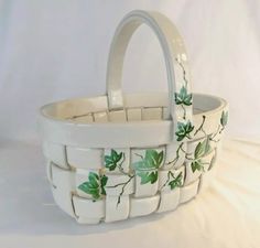 a white basket with green leaves painted on the side and handles, sitting on a white surface