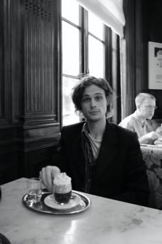 a man sitting at a table with a cup of coffee in front of him and another person behind him