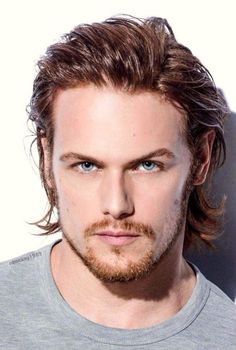 a man with long hair and blue eyes looks at the camera while wearing a grey shirt