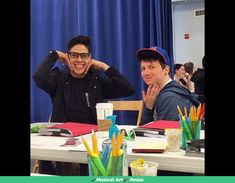 two men sitting at a table with pens and pencils in front of them smiling