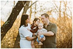 a man and two women holding a baby while standing in front of trees with leaves on the ground