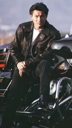 a man sitting on top of a motorcycle wearing a black leather jacket and jeans with his legs crossed