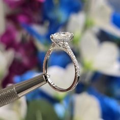 a close up of a diamond ring being held by a tweezer with flowers in the background