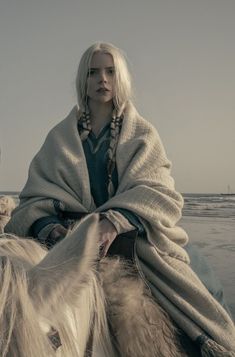 a woman riding on the back of a white horse next to the ocean with a blanket over her head