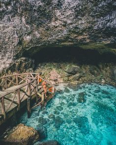 a person standing on a wooden bridge over blue water in front of a large cave