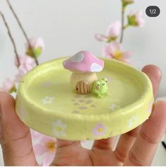 a hand holding a yellow plate with a small mushroom on it and pink flowers in the background