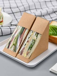 two sandwiches in a box sitting on a plate next to a sandwich wrapper with lettuce and tomato