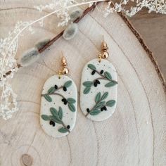 the earrings are decorated with green leaves and black berries on white marble, sitting on a piece of wood