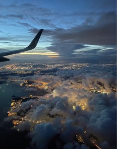 the wing of an airplane flying over clouds at night with city lights in the distance