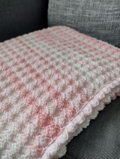 a pink and white crocheted blanket sitting on top of a gray couch next to a pillow