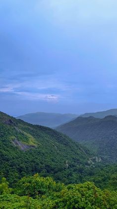 the mountains are covered with green trees and bushes in the foreground, under a cloudy blue sky